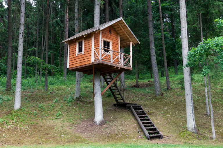 Een foto van een grote boomhut tussen de bomen. Op de voorgrond staan houten stoelen.