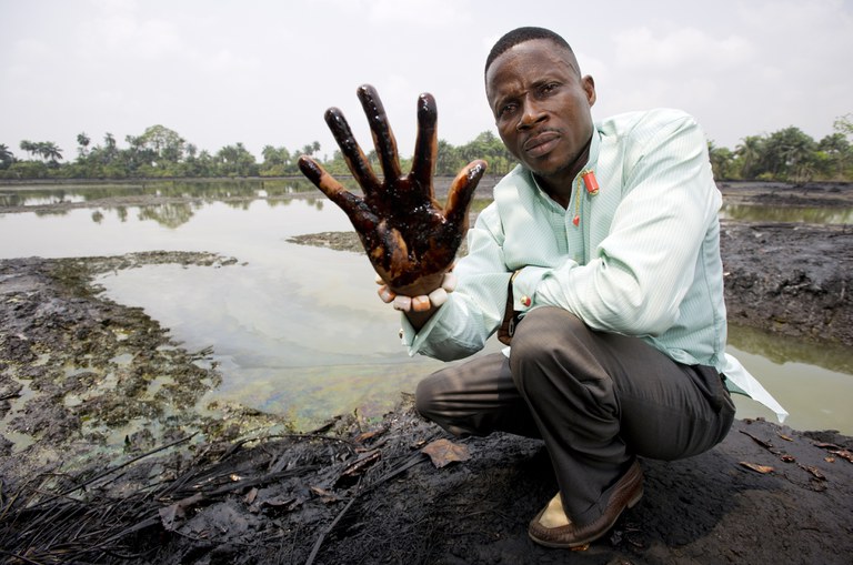 Eiser Eric Dooh toont de ruwe olie die de oevers van de kreek door zijn dorp Goi heeft aangetast. Meerdere lekkages in een pijpleiding van Shell hebben de kreek over een lengte van vele kilometers zwaar verontreinigd, waardoor vis en ander leven uit het getijdengebied zijn verdwenen.