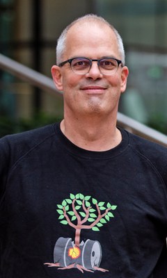 Peer de Rijk - Campaigner Team Klimaatplannen Foto van Peer de Rijk. Peer is een witte man, met grijs haar en een bril, op zijn kleding staat een tekening van een gebroken Shell-olievat waar een boom uit groeit.