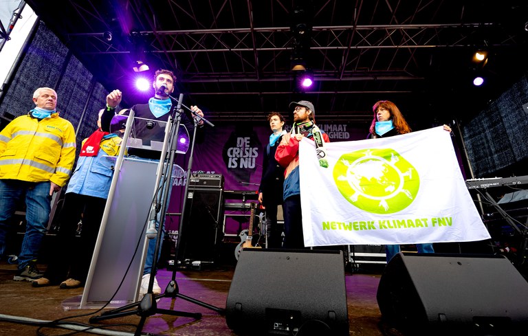 Op de afbeelding zie je een spreker van FNV tijdens de Mars van Klimaat en Rechtvaardigheid in 2023. Hij staat samen op het podium met werknemers uit de industrie en mensen van FNV Netwerk Klimaat. 