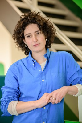 Foto van Ilana Cukier, een witte vrouw met donker krullend haar. Zij draagt een blauw overhemd. Op de achtergrond is een trap zichtbaar. 