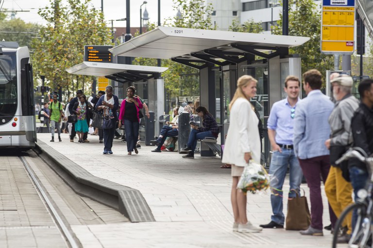 Foto van mensen op een moderne tramhalte, zo te zien bij een station. Het is er gezellig druk.