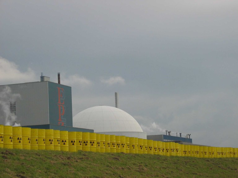 Foto van een kerncentrale met op de voorgrond een lange rij geel geschilderde stalen vaten met het symbool voor radioactiviteit.