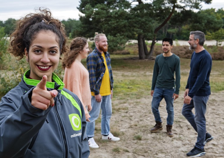 Foto van een groep jonge mensen. Op de voorgrond een lachende vrouw met een Milieudefensie-jasje aan. Ze wijst naar jou.